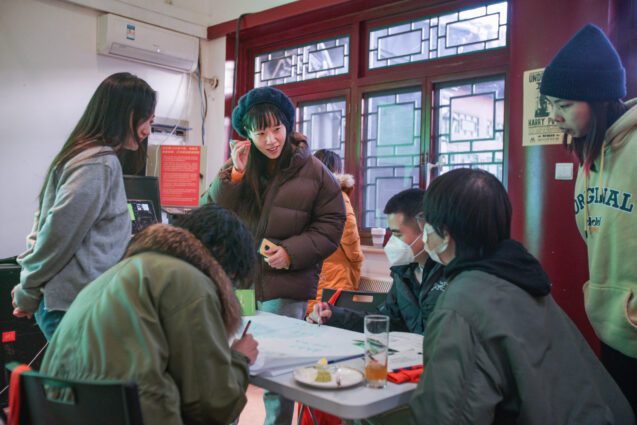 A group of Chinese people sitting and standing around a table, with a Chinese window frame in the background. One woman is standing in the centre smiling; two men are sat at the table wearing face masks