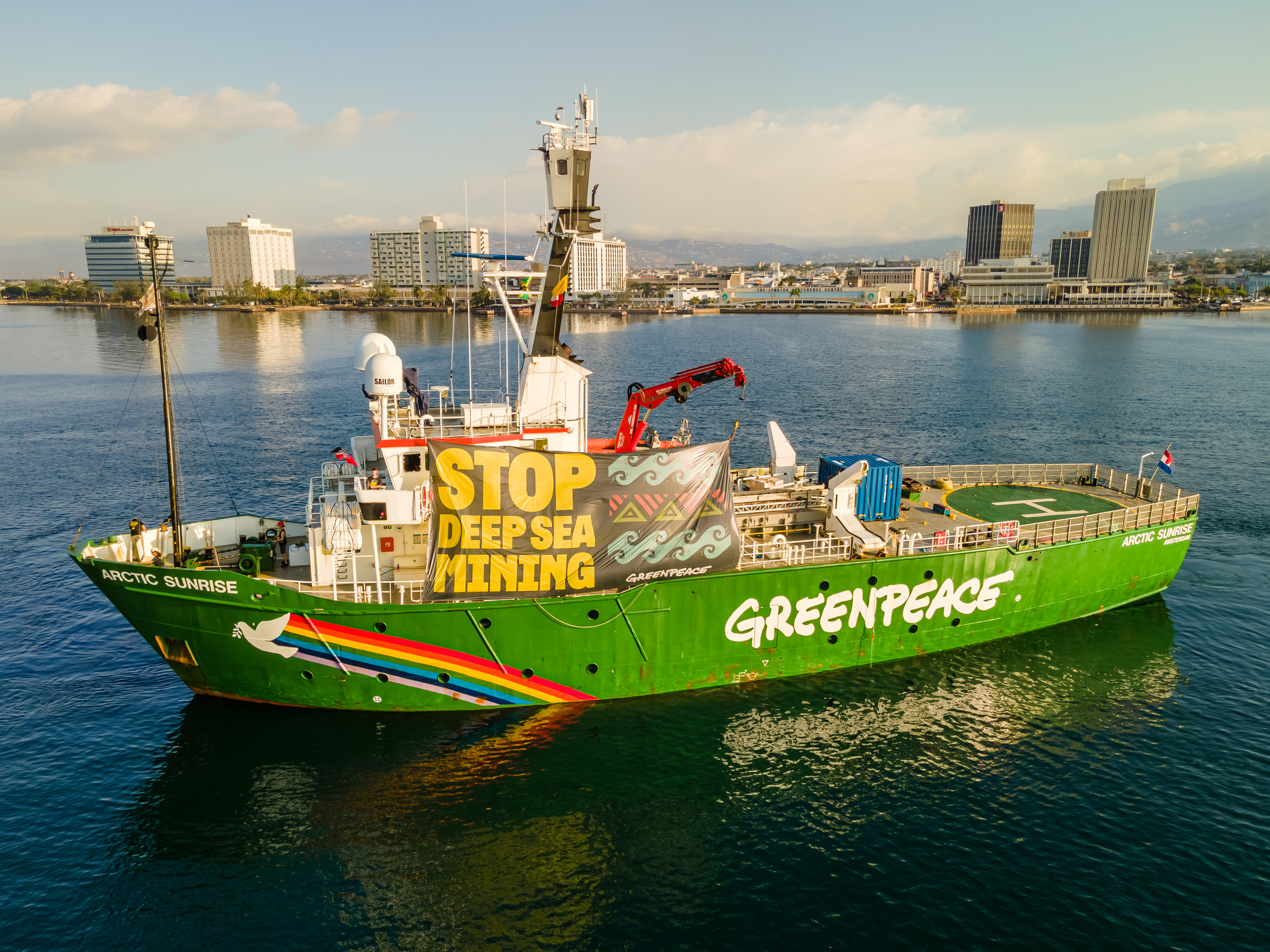 A bright green Greenpeace ship with a rainbow dove motif across the stern, sat in a still bay, with a banner hanging reading "Stop Deep Sea Mining" and a few high rise buildings in the background