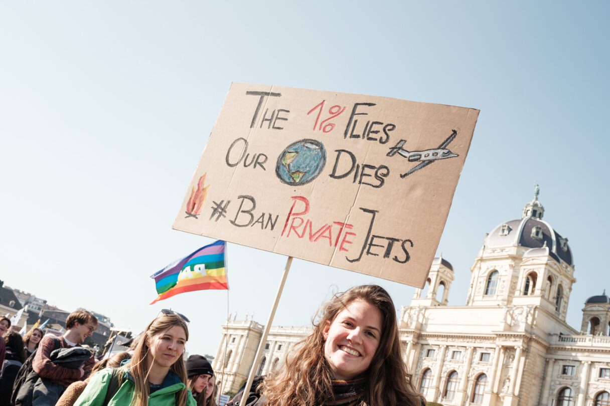 Smiling protestor holds a handwritten sign saying 'The 1% flies, our planet dies. Ban private jets.'