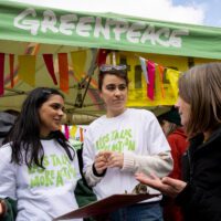 Two Greenpeace volunteers speak to a member of the public in front of a Greenpeace branded marquee decorated with bunting.