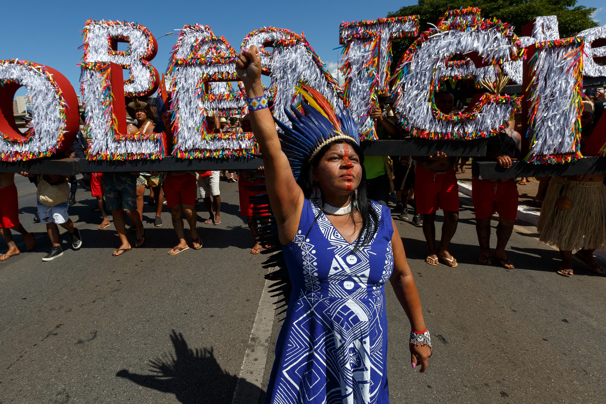 A woman with a blue yellow and red feather headdress and blue patterned dress stands on a road with her arm held high, in front of a colourful and glittery display of large lettering under which the legs of the people holding them can be seen