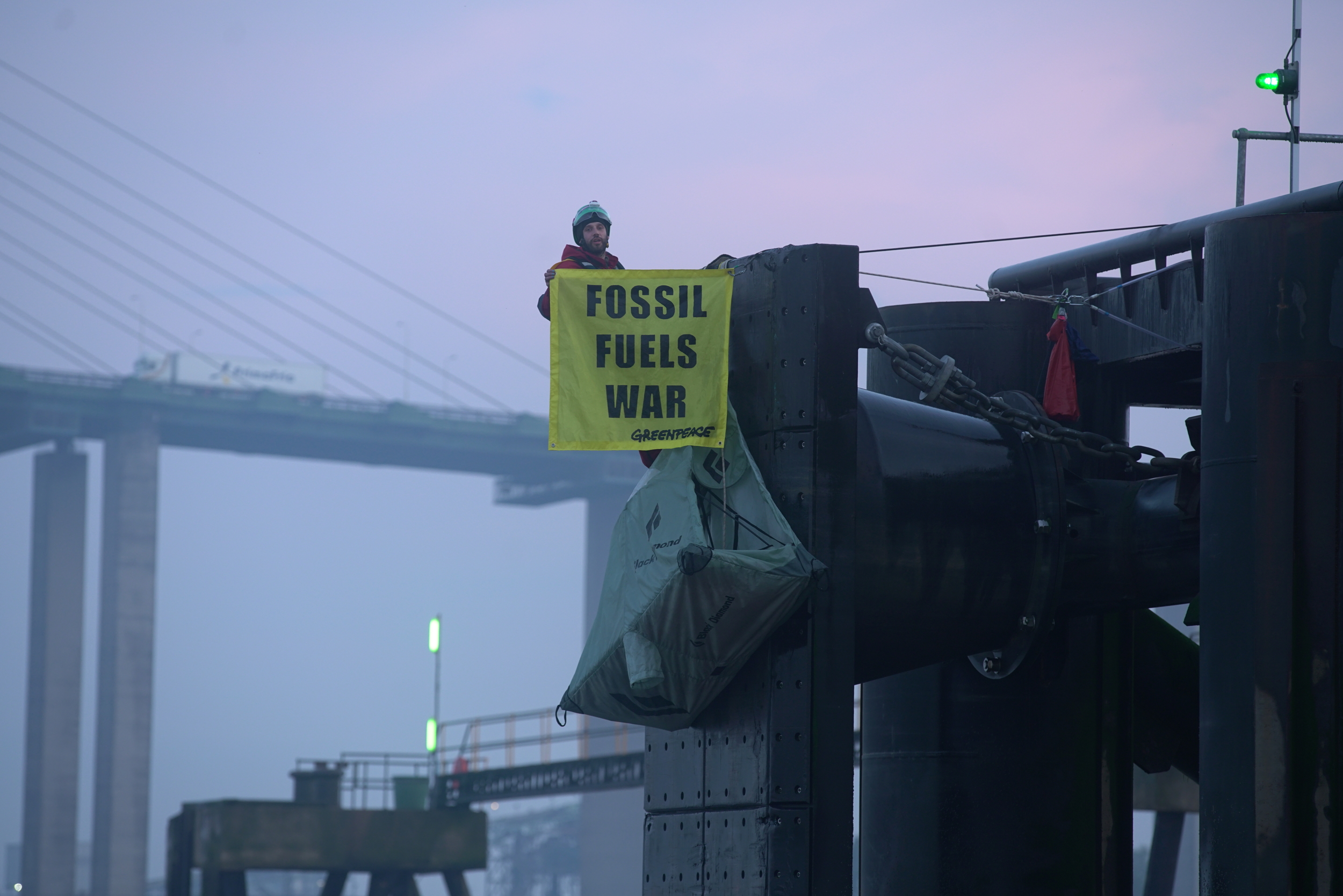 A climber hanging on a large metal structure holds a yellow banner reader 'Fossil Fuels War', in the misty background there is a suspension bridge.