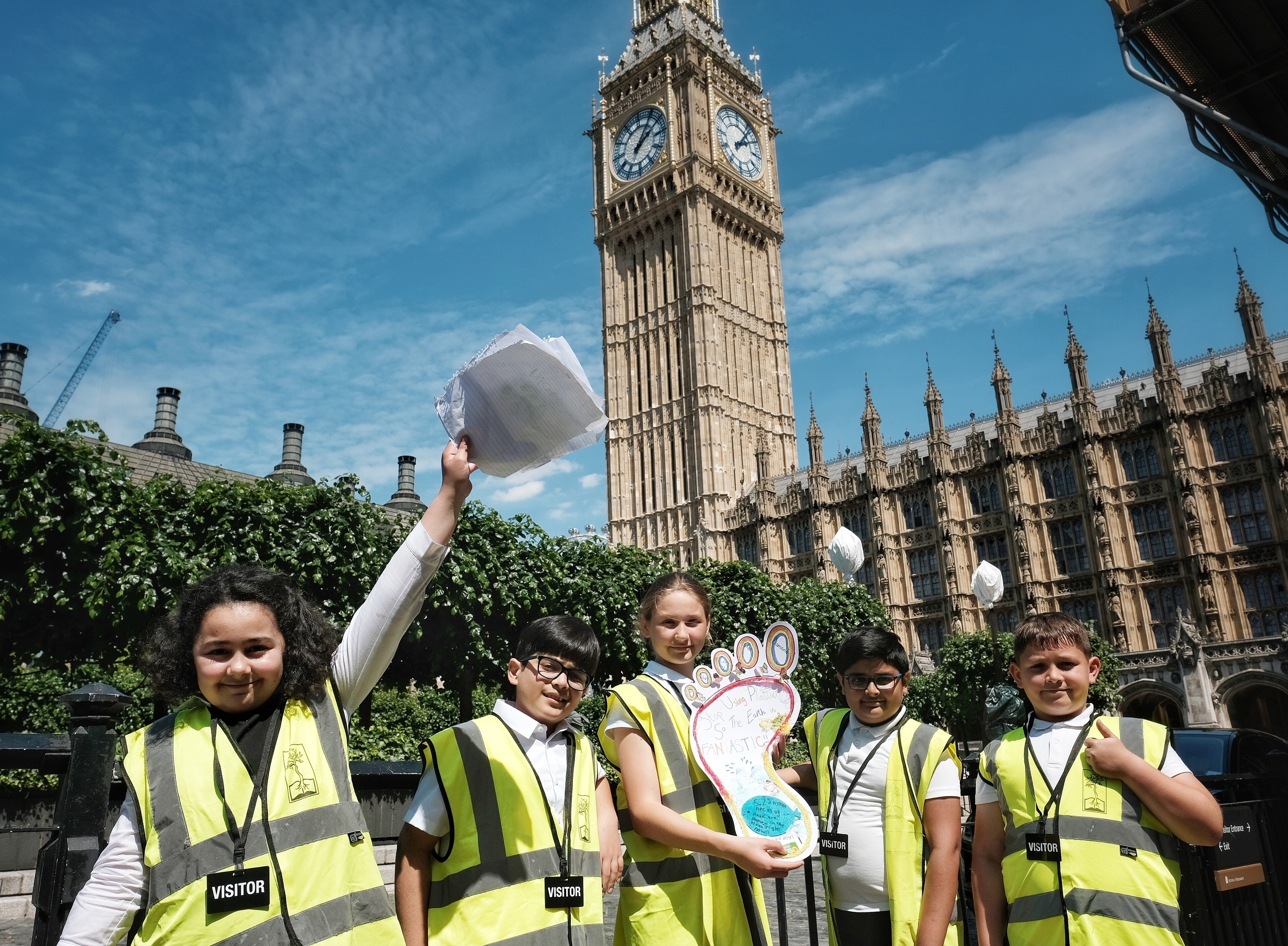 In front of a blue sky and Big Ben, five schoolchildren stand in yellow high viz jackets and 'visitor' lanyards stand. One is holding up pieces of lined paper high with her hand, another is holding a foot shape with children's writing on it