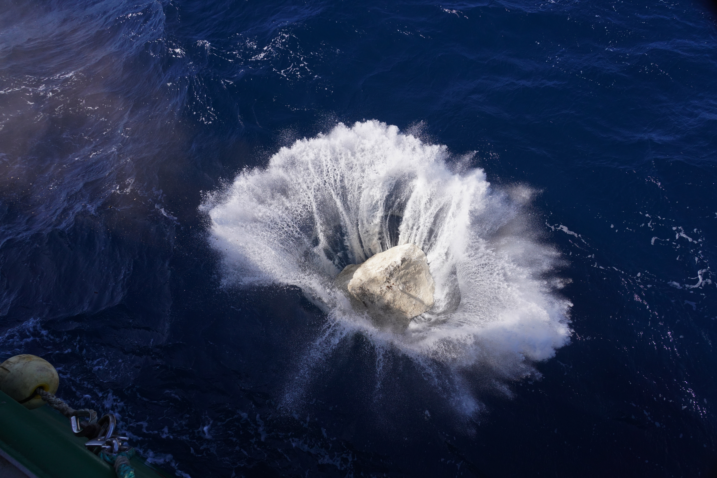 The moment a large rock splashes into deep blue water, with a large spray as the boulder is last seen before it submerges