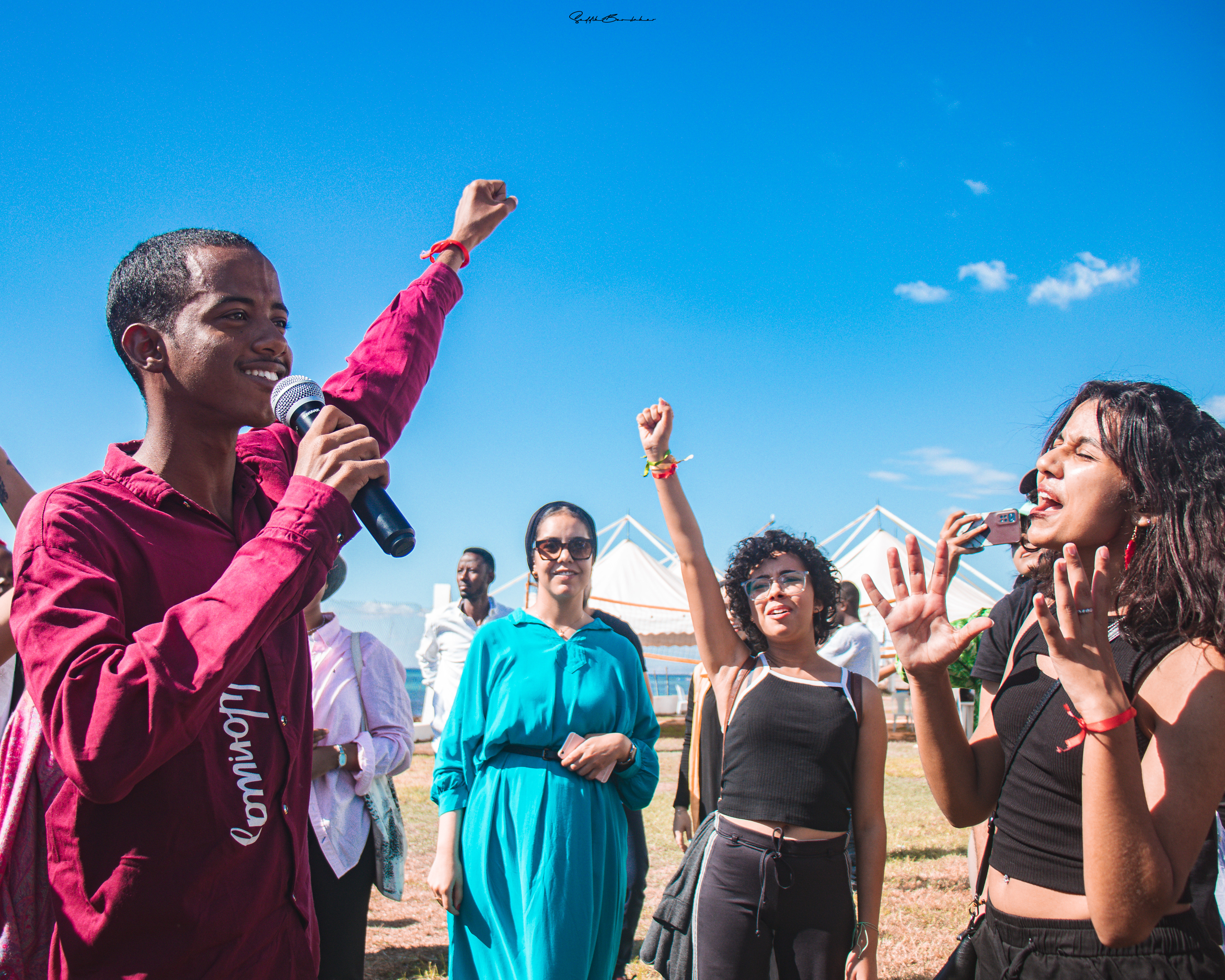 Against a blue sky a smiling dark-skinned young man holds his fist in the air and a microphone. Another young girl is central, also with her fist in the air, and another young woman holds both her hands up in front of her face, which has a scrunched expression as if she is shouting. There are more people of colour visible in the background, where there are white tents and a sliver of sea.