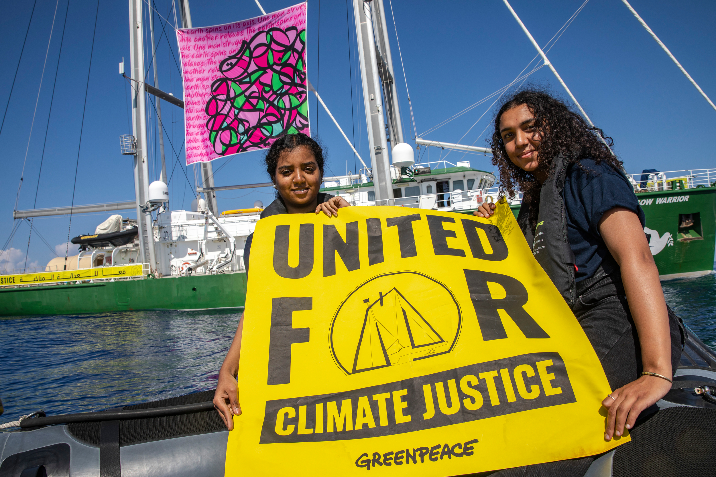 Two young women hold a large yellow Greenpeace banner reading 'United for Climate Justice'. In the background a green and white ship is visible which has a colourful pink and green banner hanging on its sails.
