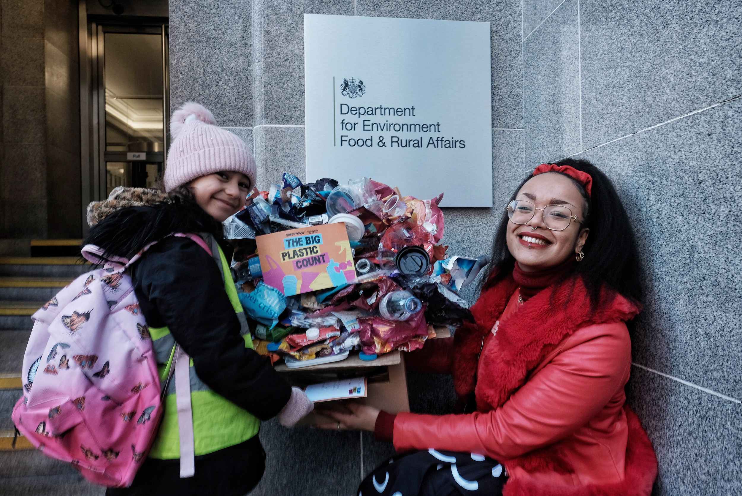 A woman in red with long black hair and glasses smiles with young girl in a high viz with a ptin bobble hat and backpack. They are holding a box piles high and overflowing with plastic rubbish, with a flyer on it reading 'The Big PLastic Count'. They are stading in front of the Department for Environment Food and Rural Affairs