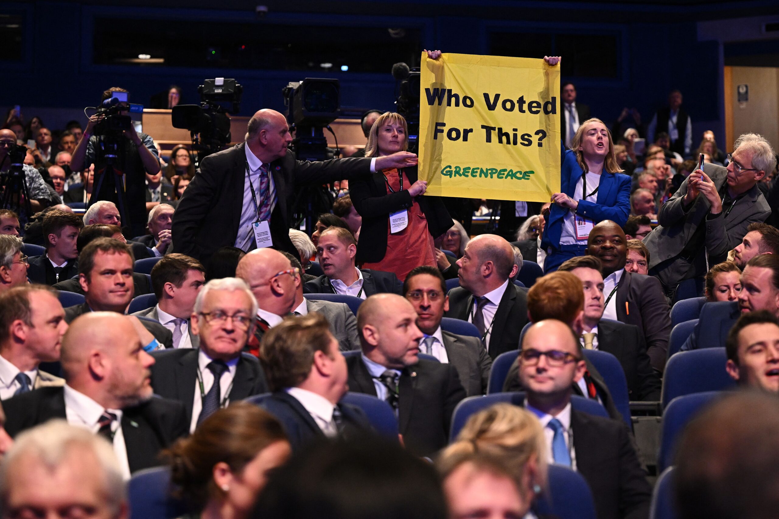 A crowded theatre full of predominantly white men in suits. Two women standing wearing lanyards are holding a yellow Greenpeace banner reading 'Who Voted For This' as a man in a lanyard reaches over to grab it, and another man takes a photo from the side. In the background are large TV cameras