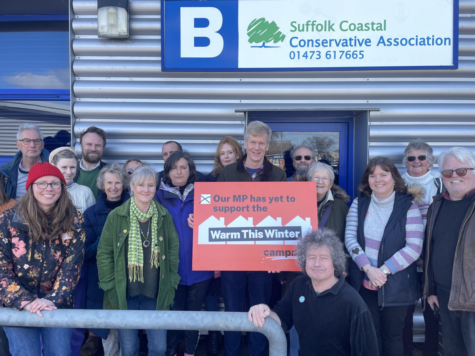 group of 17 people, mostly older and white, stand in front of. metal clad building with a sign reading "Suffolk Coastal Conservative Association 01473617665". In the middle one man holds a red right reading "Our MP has yet to support the Warm This Winter campaign"