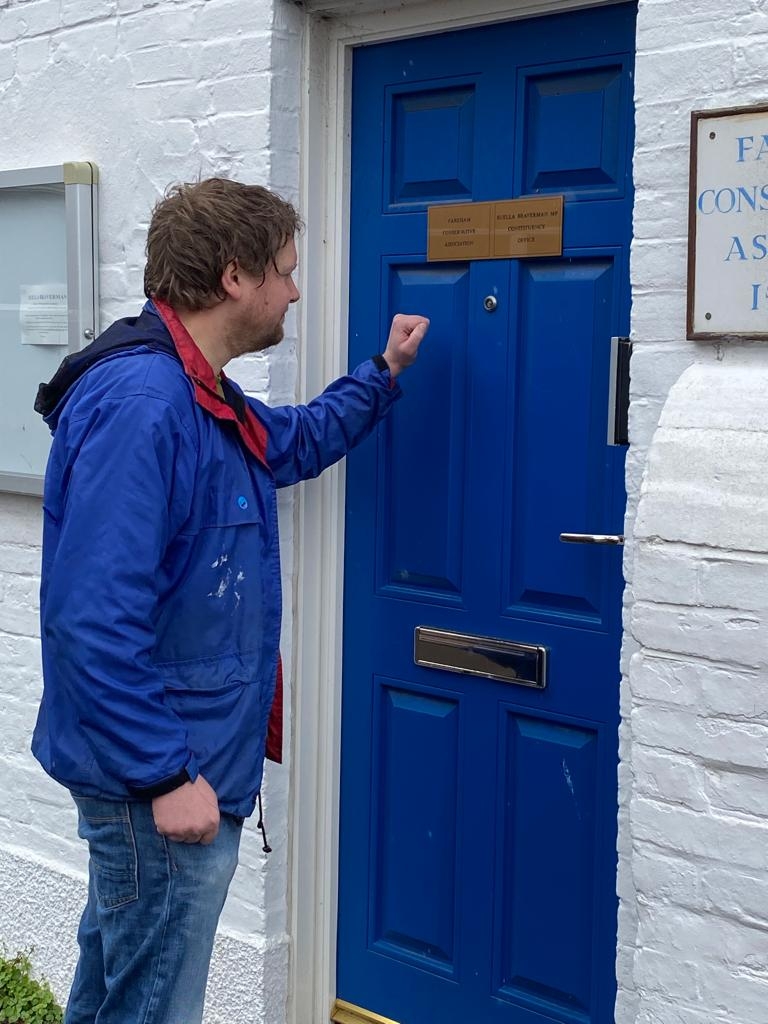 A man in a blue jacket approaches a blue door with his arm up as if he is about to knock on it. There is a sign partly visible next to the door with lettering suggesting it is an MP's office