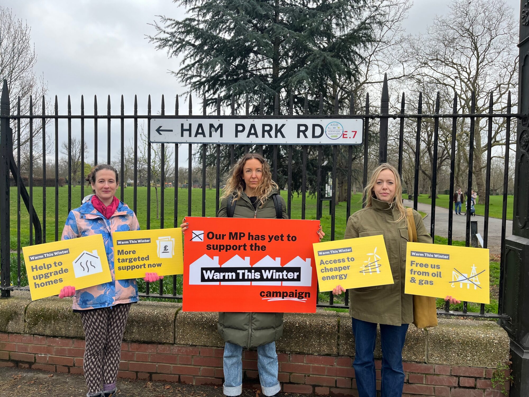 Three women stand in front of a park fence, on which is a road sign reading "Ham Park Rd, E7". They are dressed in warm coats and holding signs, small ones in yellow on the side and a larger red one in the middle. The yellow signs say "Help to upgrade homes" and "More targeted support now" on one side and "Access to cheaper energy" and "Free us from oil and gas" on the other. The red sign in the middle reads "Our MP has yet to support the Warm This Winter campaign". All signs include stylised icons and graphics of houses, money, windfarms and oil and gas drilling