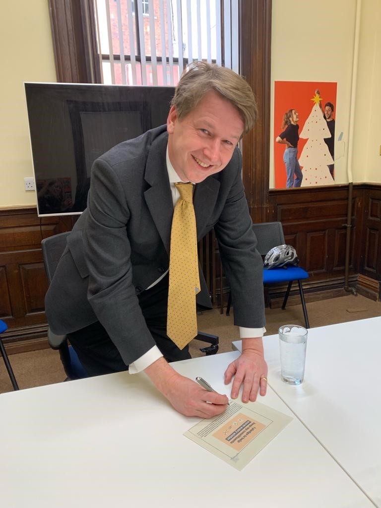 A smiling man in a dark suit and yellow tie leans standing over a table, about to sign a piece of paper.