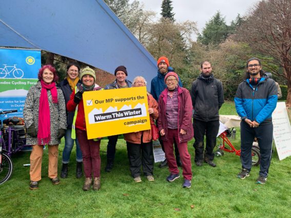 Nine people of mixed genders and races stand on grass in front of a tent with a sign next to it showing a bicycle icon. Two at the front hold a yellow sign reading "Our MP supports the Warm This Winter campaign"