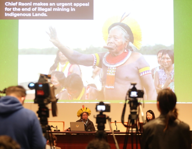 A forest of cameras are trained on an indigenous leader in traditional headwear giving a speech.