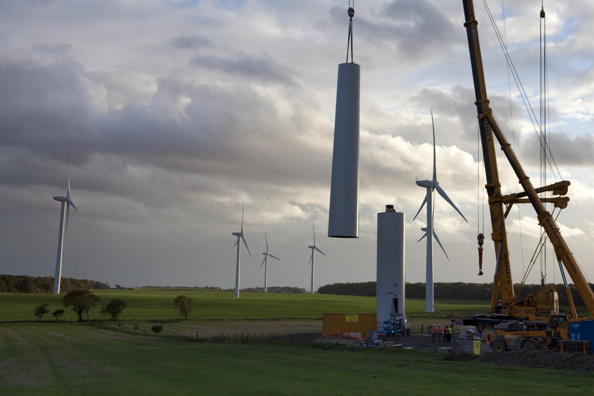 Giant cranes assemble the tower of a new wind turbine. A dramatic sunny sky and completed turbines can be seen in the background.