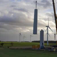 Giant cranes assemble the tower of a new wind turbine. A dramatic sunny sky and completed turbines can be seen in the background.