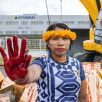 An Indigenous woman in traditional dress holds up a red-stained hand to the camera. In the background there's a Hyundai-branded building and an inflatable digger covered in red handprints.