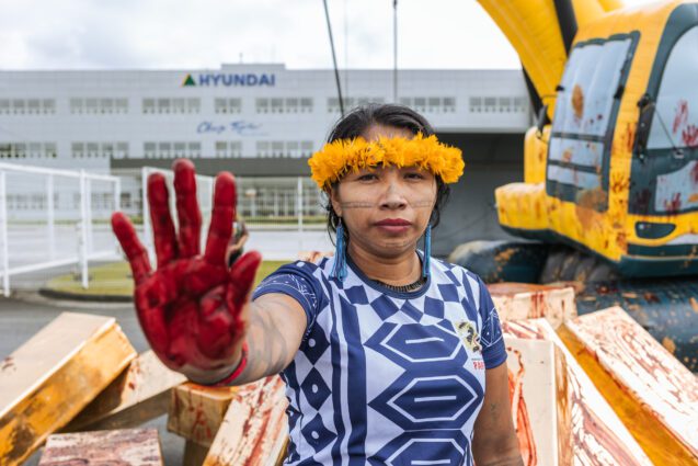 An Indigenous woman in traditional dress holds up a red-stained hand to the camera. In the background there's a Hyundai-branded building and an inflatable digger covered in red handprints.