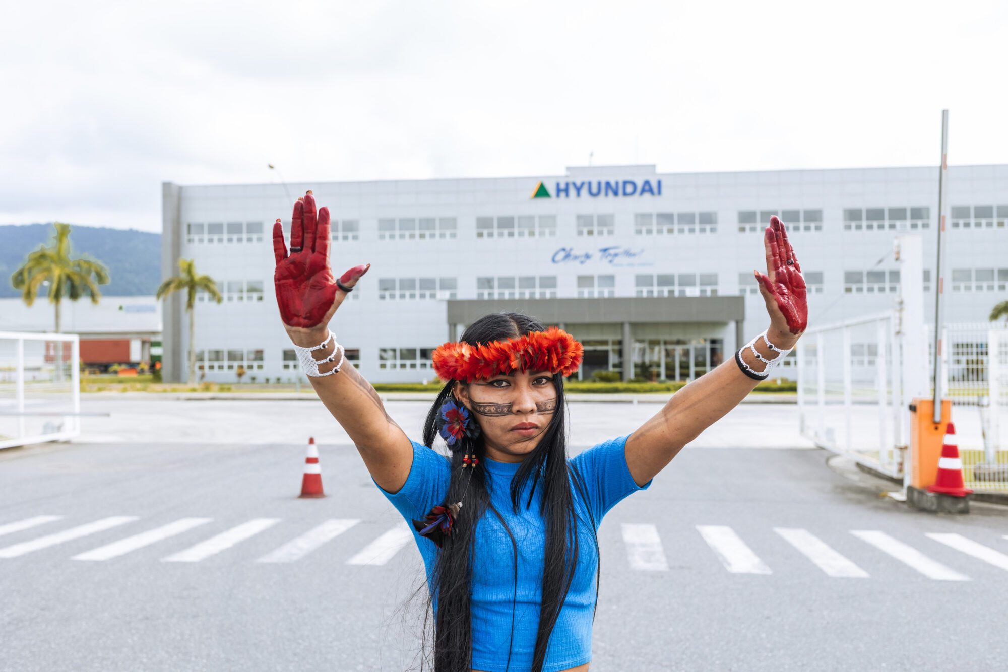 An indigeous activist in traditional headwear holds up red-stained hands to the camera. In the background there's a factory building with the Hyundai logo on the front.
