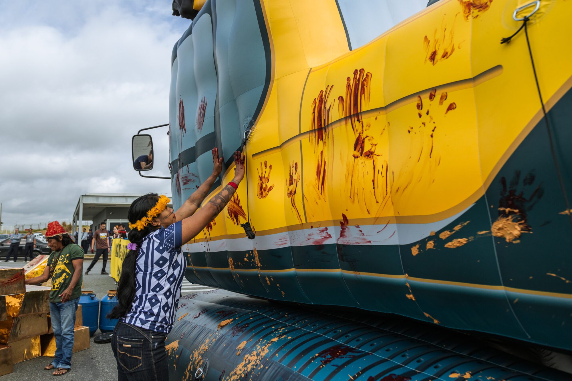 Indigenous activists in traditional headwear mark the side of a yellow excavator with red handprints.