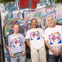 Three people wearing tshirts with the same colourful design stand in front of a wall covered in graffiti. They're posing informally and smiling into the camera.