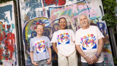 Three people wearing tshirts with the same colourful design stand in front of a wall covered in graffiti. They're posing informally and smiling into the camera.