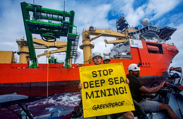 Greenpeace activists on a small boat hold up a stop deep sea mining banner in front of a giant industrial ship