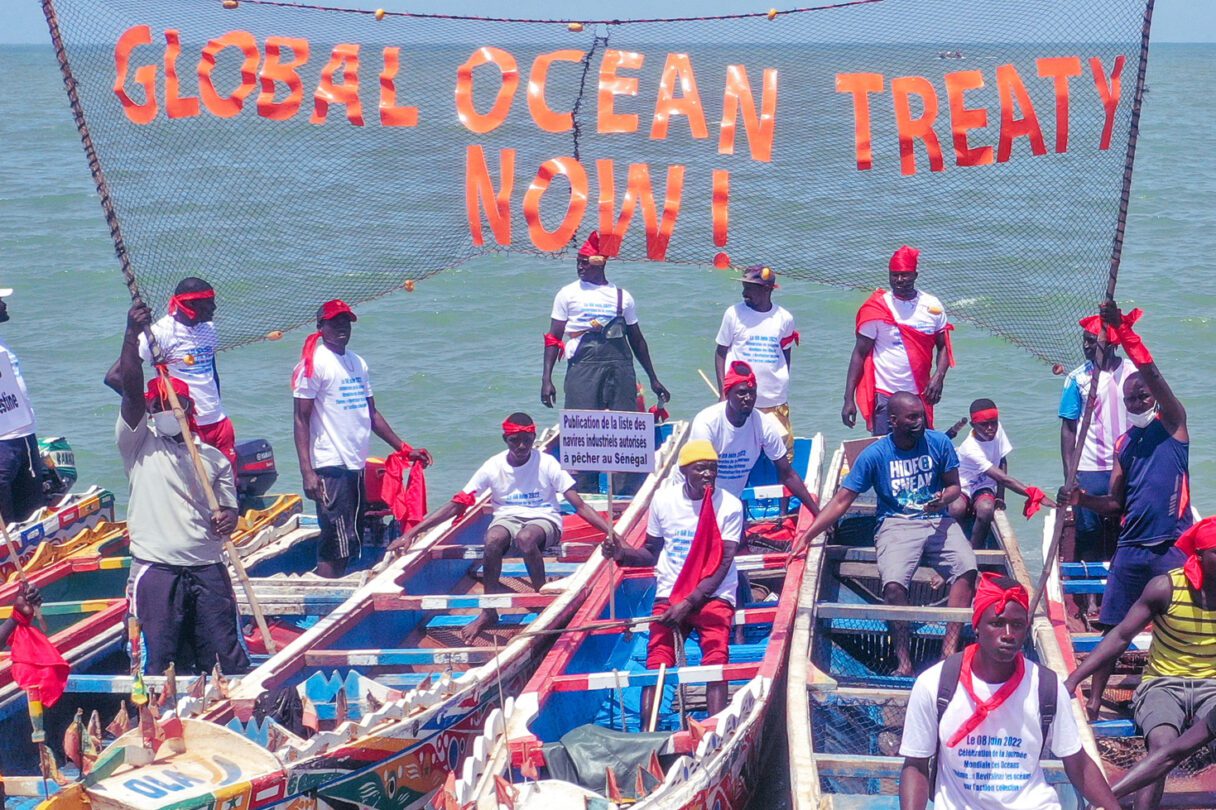 People stand on brightly painted traditional Senegalese fishing boats. A banner between the boats reads Global Ocean Treaty Now.