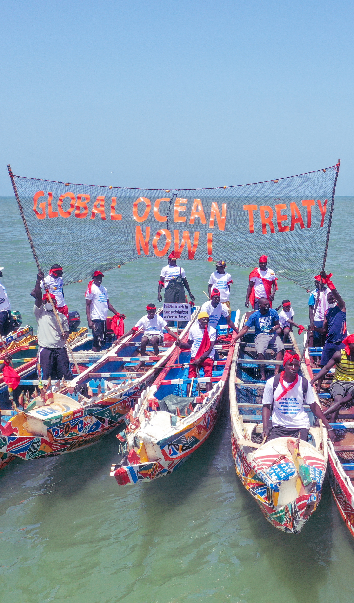 People stand on brightly painted traditional Senegalese fishing boats. A banner between the boats reads Global Ocean Treaty Now.