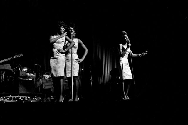 Black and white photo of three Black women standing on a stage in matching dresses, singing into microphones.