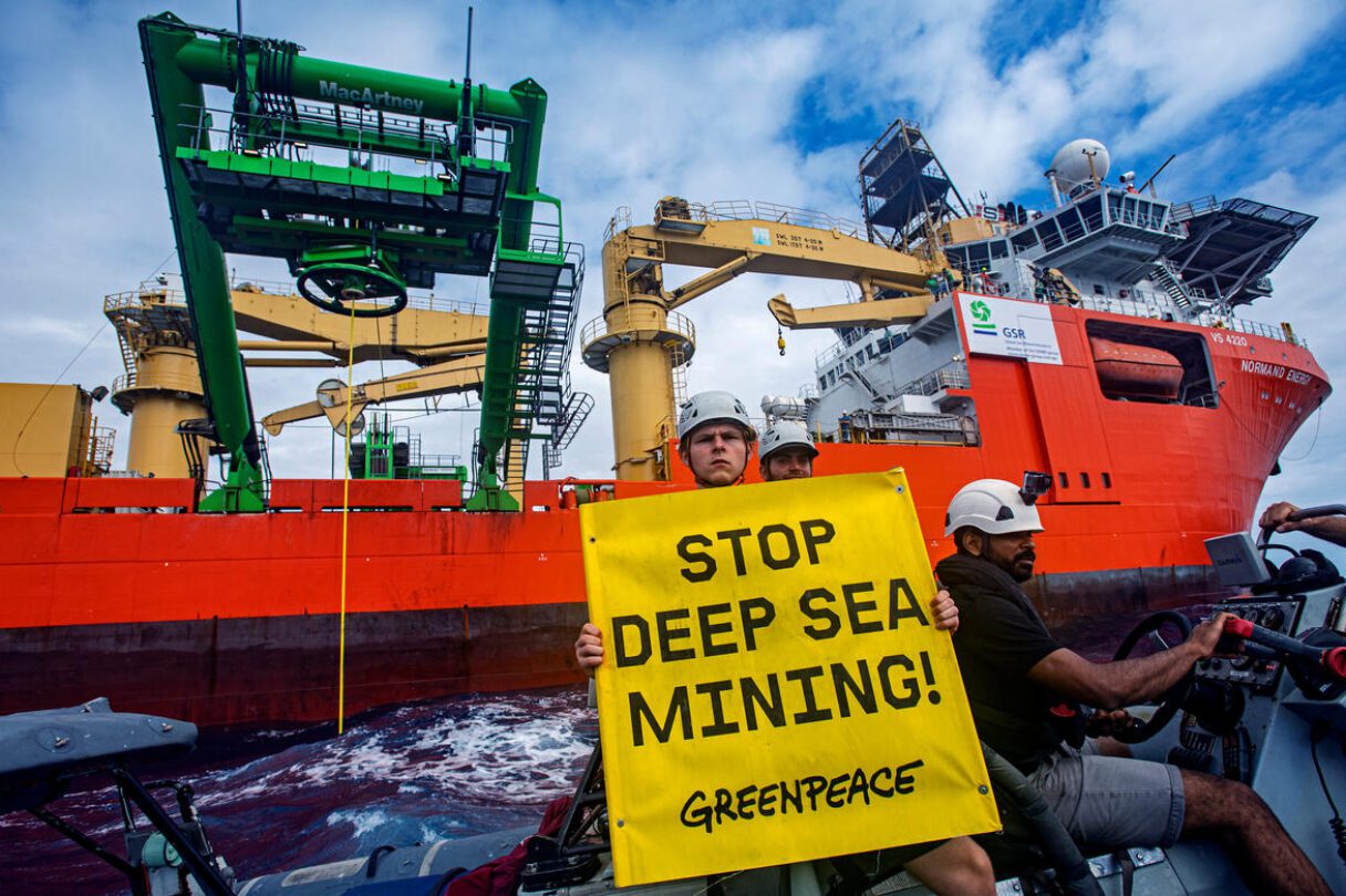 Greenpeace activists on a small boat hold up a stop deep sea mining banner in front of a giant industrial ship