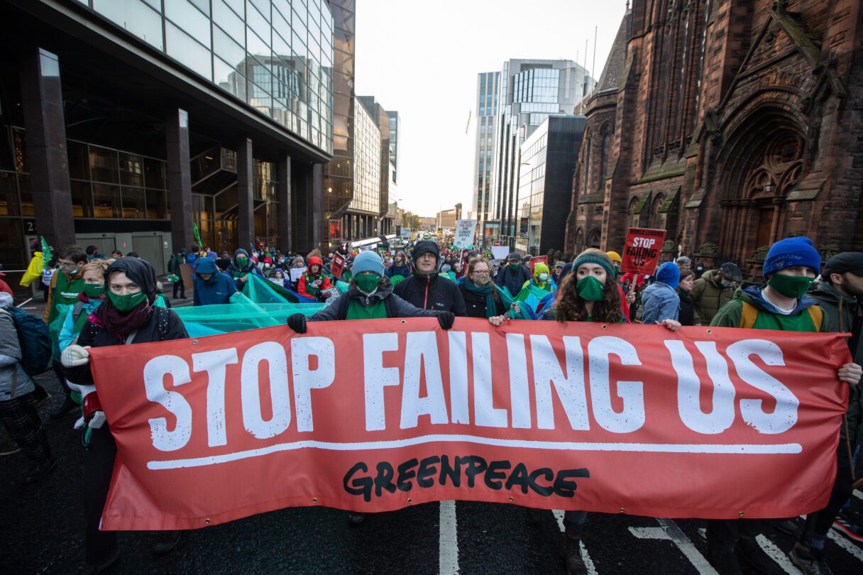 Protesters march through a city street of new glass and old gothic buildings. They are wearing green facemasks and holding a massive red banner that reads: STOP FAILING US and Greenpeace
