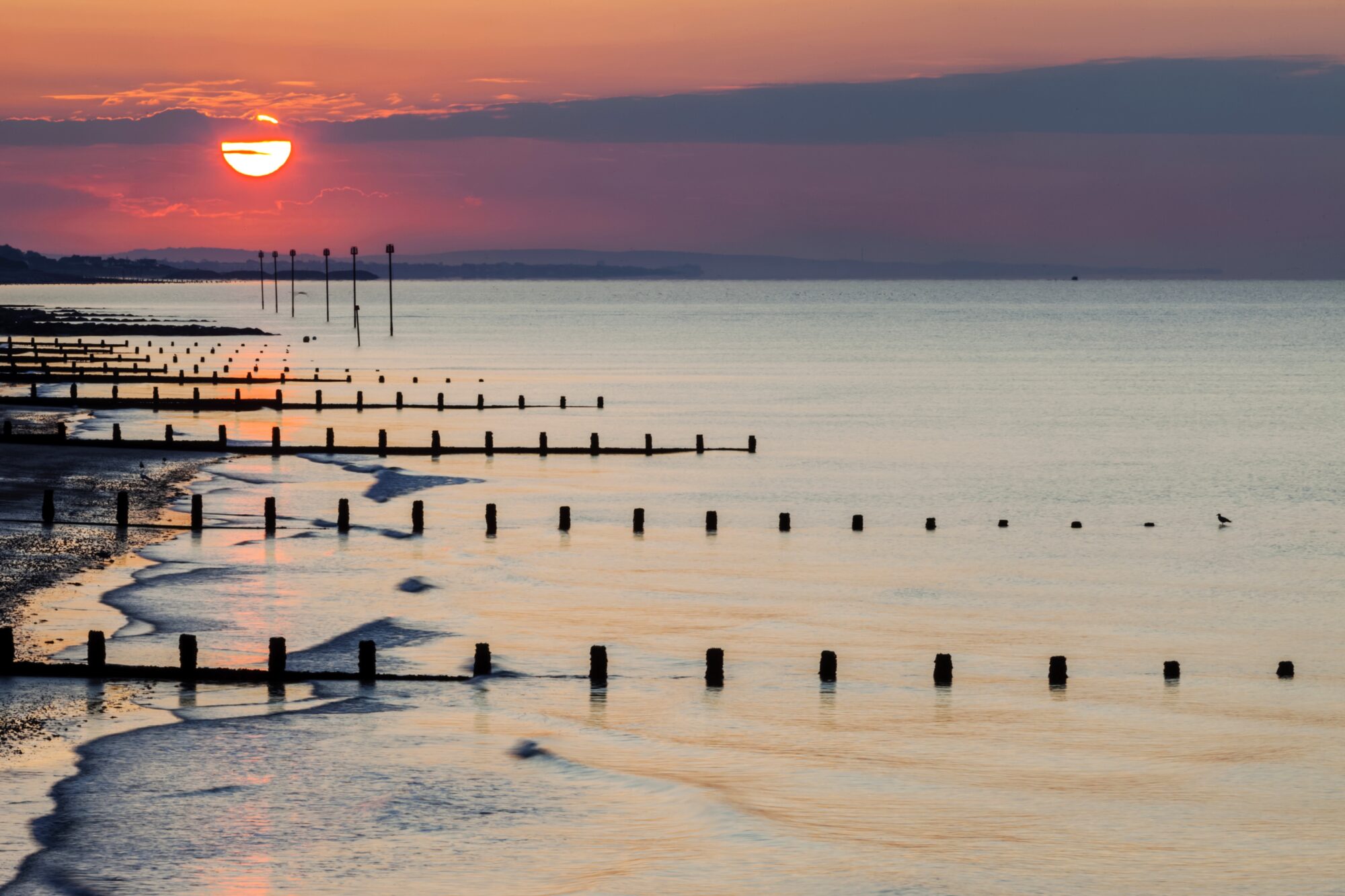 A glowing red sunrise through purple and orange clouds, over a still seascape and coastline emerging from the left with wooden groynes stretching out into the sea in perpendicular lines along the whole coast, silhouetted by the sunrise