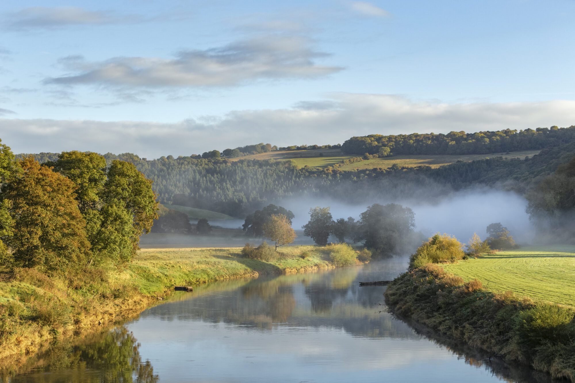 A landscape of the river wye, with green and tyellow pasture and trees stretching into the distance. In the foreground a wide river, which melts back into a misty glade among trees and a cloud-spotted pale blue sky