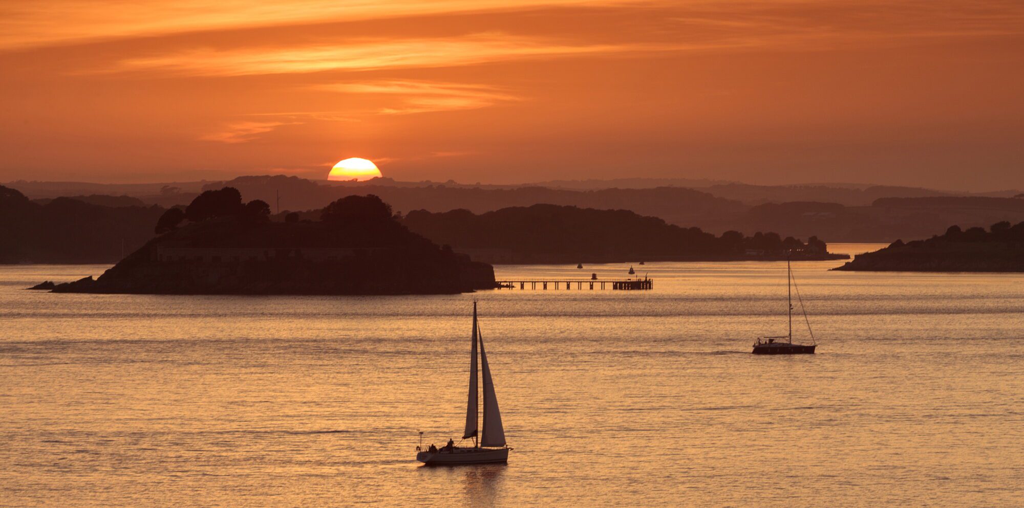A bright orange sunset sky with a half-sun glowing orb visible just behind mountains and small estuary islands. A central island has a jetty silhouetted by the light. In the foreground, two sailboats, one with a sail up and one without, are also silhouetted on the still water.