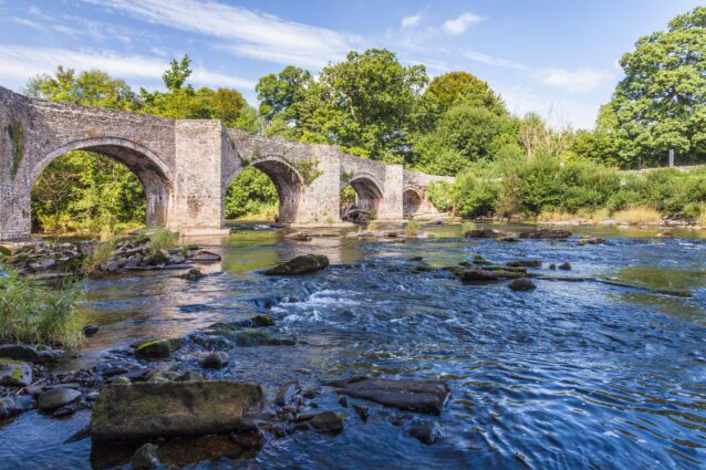 A beautiful ancient looking multi-arched stone bridge over a rocky shallow river, showing some areas of fast-flowing water, with bright green trees all around