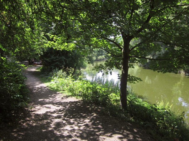 A riverside scene covered in trees and greenery, with sun and shade dappling a path that leads around the riverside bend. An inflatable is being carried in the distance on the path.