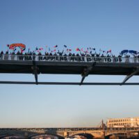 A close up of a part of a city bridge covered in people, against a blue sky, another bridge and and some buildings. The people on the bridge carry red and blue flags. A large banner central in the frame, reading "What comes next is up to us". smaller banners either side in red and blue read "Open minds" and "Open hearts"