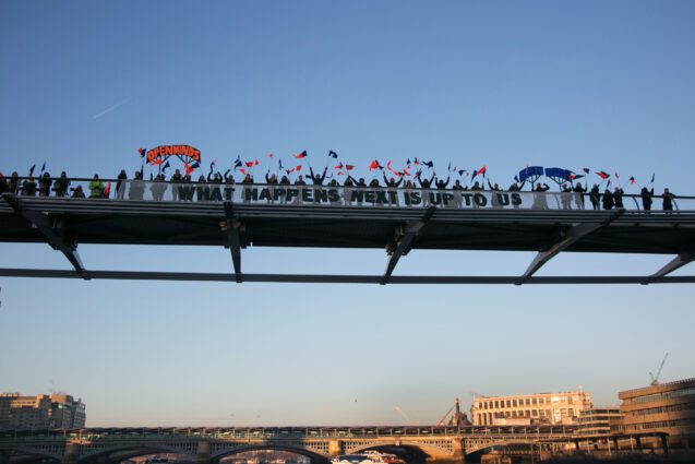 A close up of a part of a city bridge covered in people, against a blue sky, another bridge and and some buildings. The people on the bridge carry red and blue flags. A large banner central in the frame, reading "What comes next is up to us". smaller banners either side in red and blue read "Open minds" and "Open hearts"