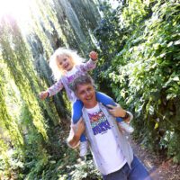 An adult carries a child on his shoulders. Sunlight shines through the trees in the background and illuminates the child's blonde hair.