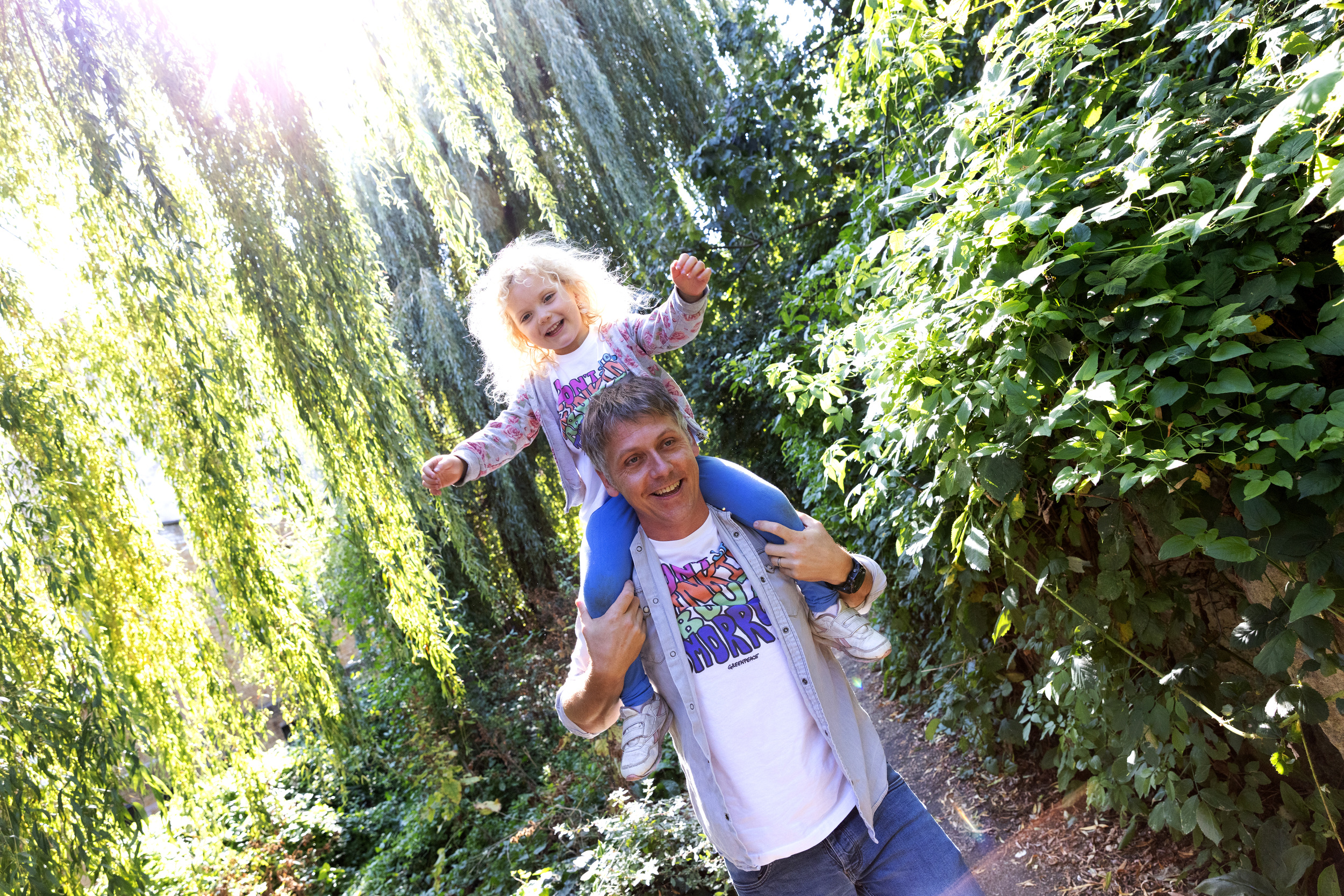 An adult carries a child on his shoulders. Sunlight shines through the trees in the background and illuminates the child's blonde hair.