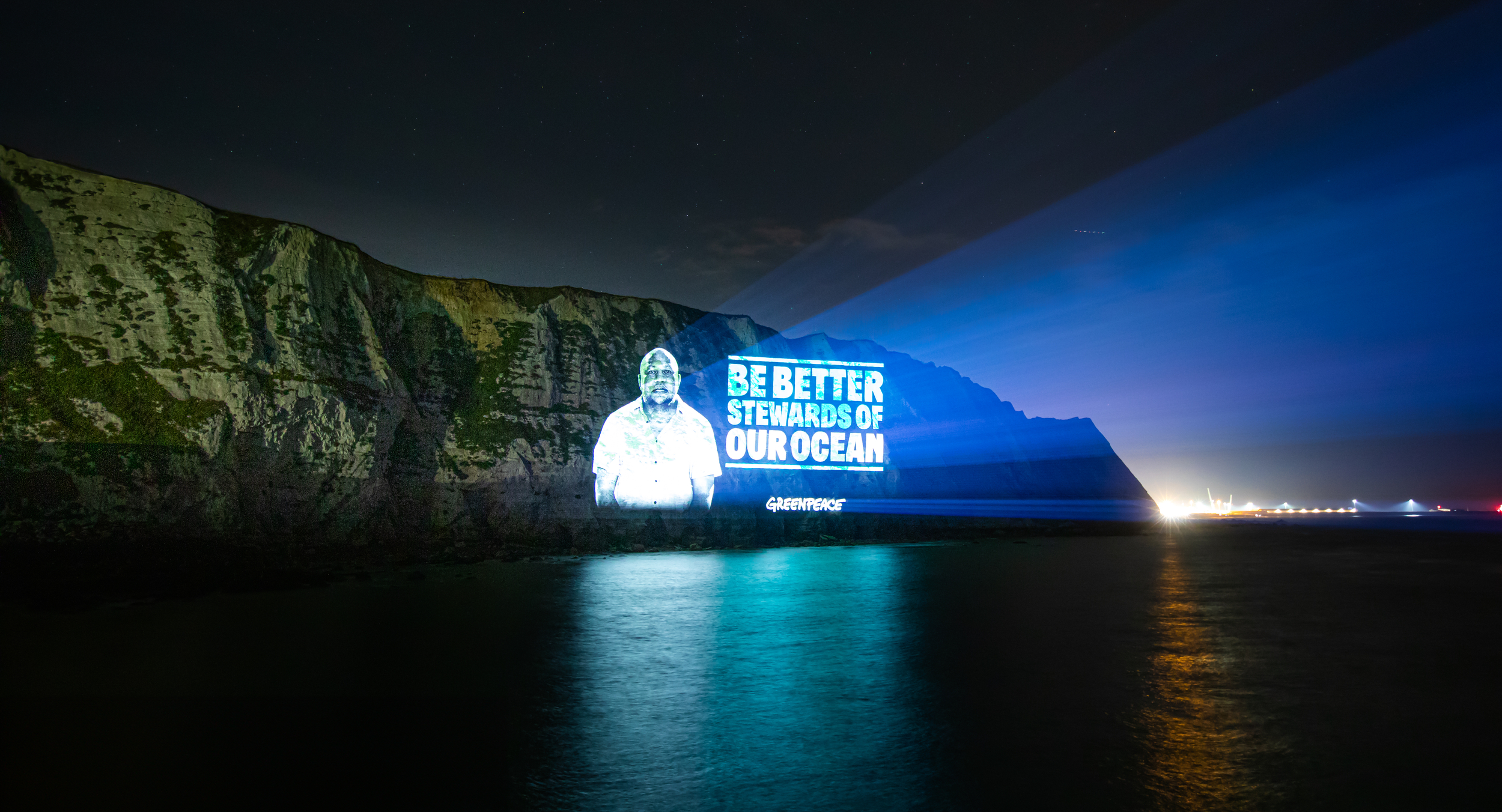 Light beams onto a cliff face at night, revealing a portrait next to the words 'Be better stewards of our ocean'