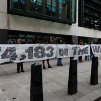 A 30 metre banner is held open by volunteers on a pavement outside an office. The banner reads "We, 174,183 on the waiting list demand allotments". The black text is grainy from a distance, as the banner is made from seed paper.