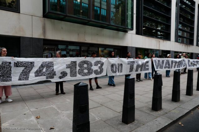 A 30 metre banner is held open by volunteers on a pavement outside an office. The banner reads "We, 174,183 on the waiting list demand allotments". The black text is grainy from a distance, as the banner is made from seed paper.