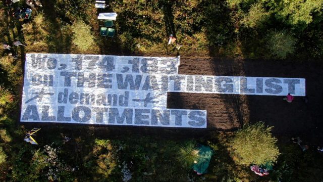 Aerial photo. A giant seed paper banner lies on bare earth ready to sprout. The banner reads "We, 174,183 on the waiting list demand allotments".