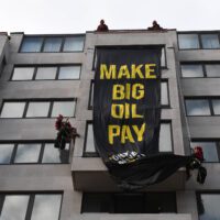 Climbers on a building unveil a big black banner with yellow text. The banner reads: Make big oil pay