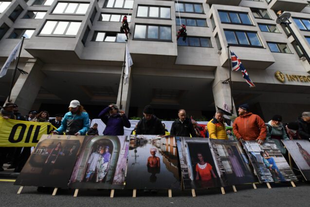 Protesters stand outside, behind printed portraits of people in flooded streets and homes.