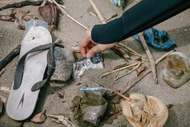 A hand reaches down to pick up a Dove-branded plastic sachet that's half buried in beach sand. A broken sandal is also visible in frame.