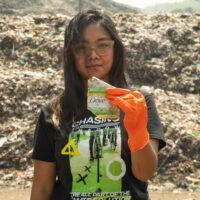 An activist in a Greenpeace t-shirt holds up a piece of plastic litter with Dove branding. There's a huge pile of plastic waste in the background.