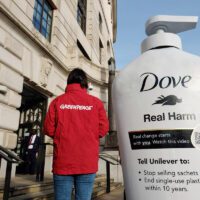 A giant Dove pump bottle stands in front of a white London building. A Greenpeace activist in a red jacket stands next to it, and it towers over her at around twice her height. The bottle reads "Dove Real Harm" "Real Change starts with YOU. Watch this video" with a QR code. It also reads "Tell Unilever to: Stop selling sachets now. End single use plastic within 10 years"